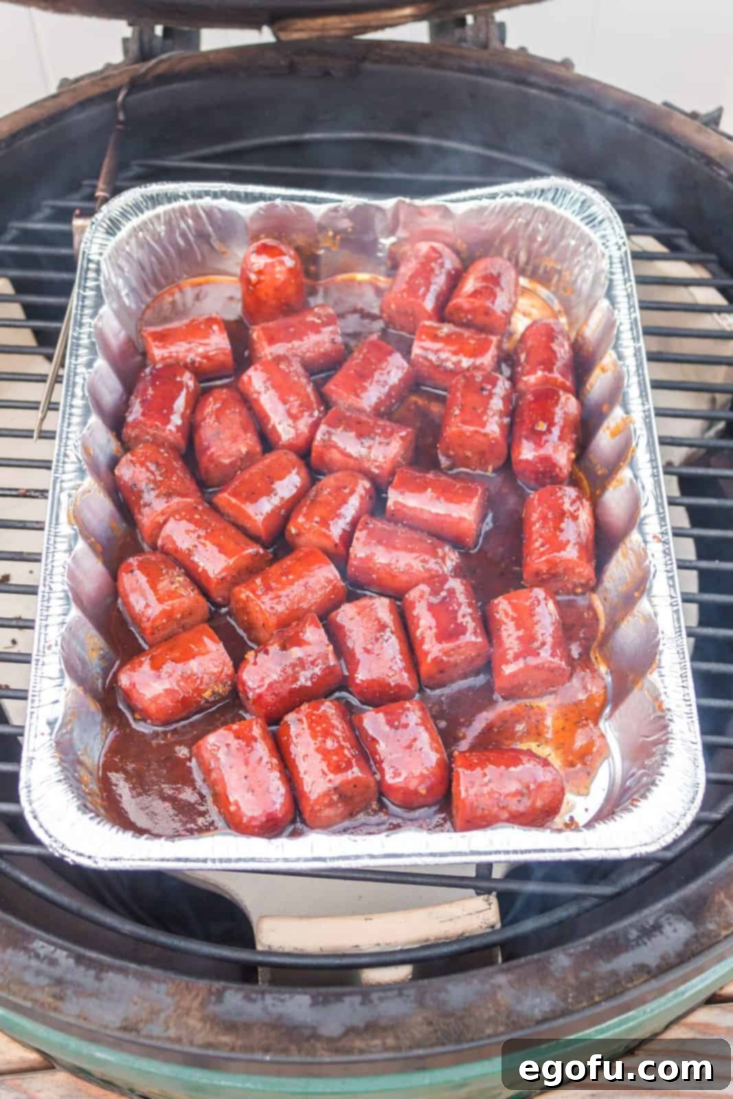 Cut up hot dogs with seasoning and sauce in an aluminum pan on a grill.