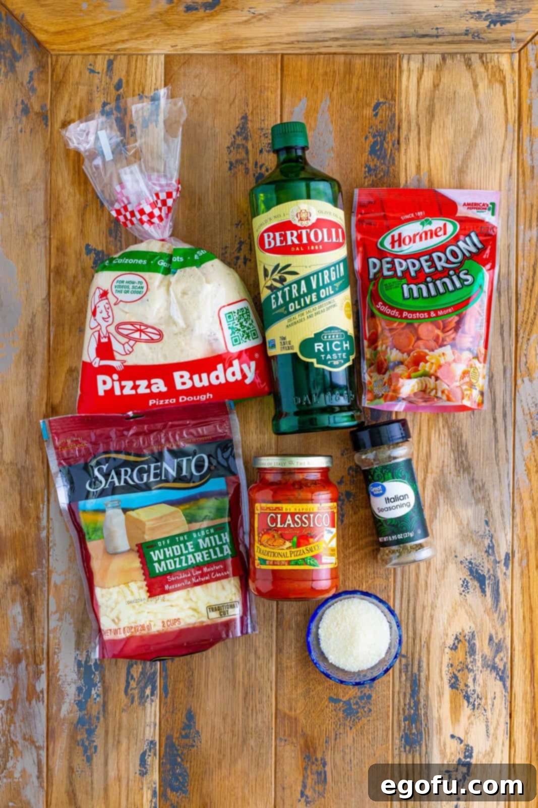 A vibrant flat lay of all the essential ingredients: pizza dough, pepperoni, a bottle of olive oil, mozzarella cheese, pizza sauce, Italian seasoning, and parmesan cheese.