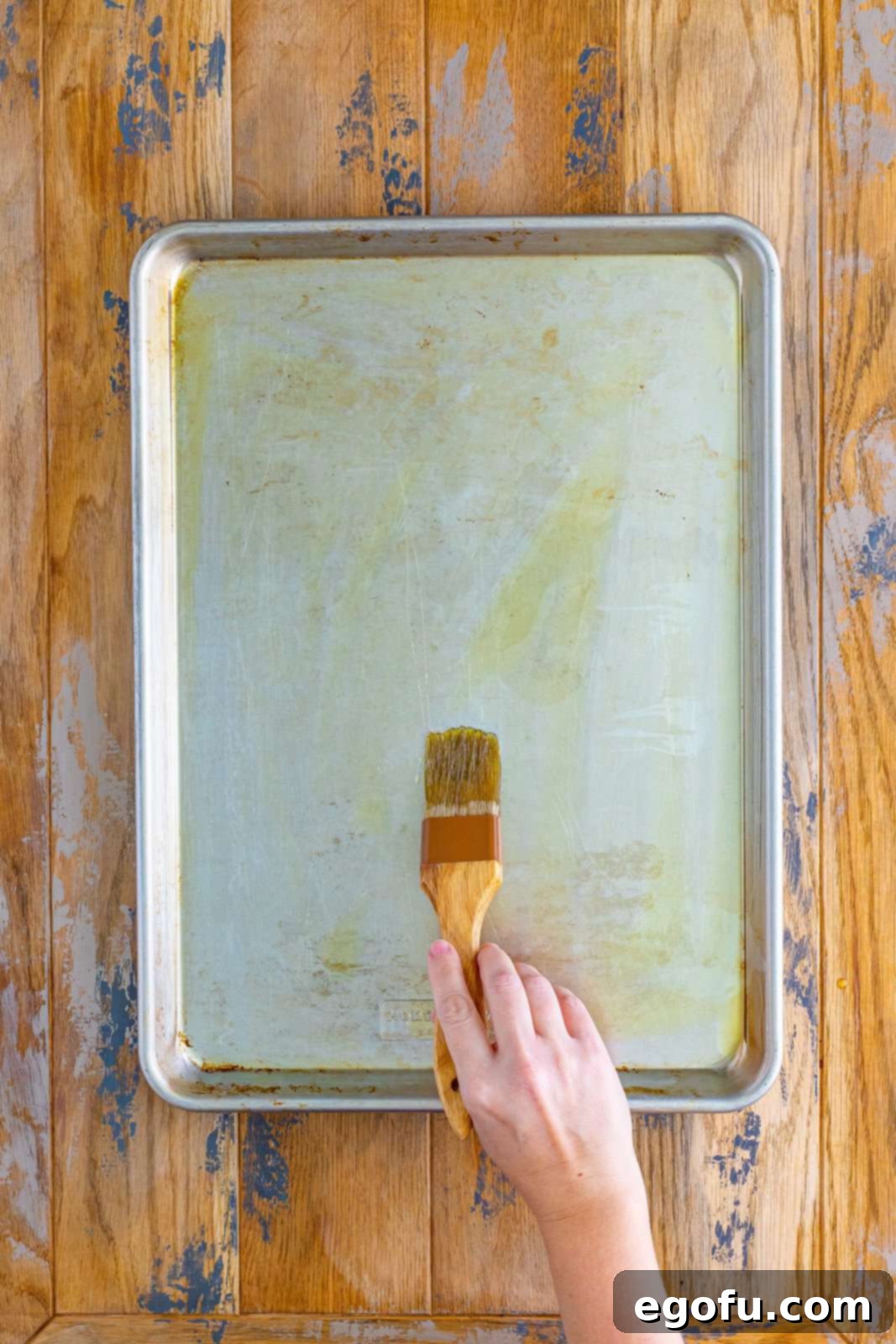 A hand expertly brushing olive oil evenly across a metal sheet pan, preparing it for the pizza dough.