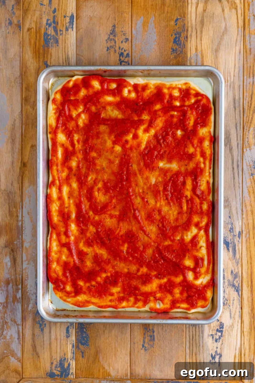 Rich red pizza sauce generously spread across the pre-baked pizza dough, covering it uniformly to the edges.