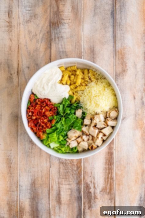 A bowl of rotini pasta, diced grilled chicken, bacon, romaine lettuce, shredded parmesan cheese, and Caesar dressing before mixing.