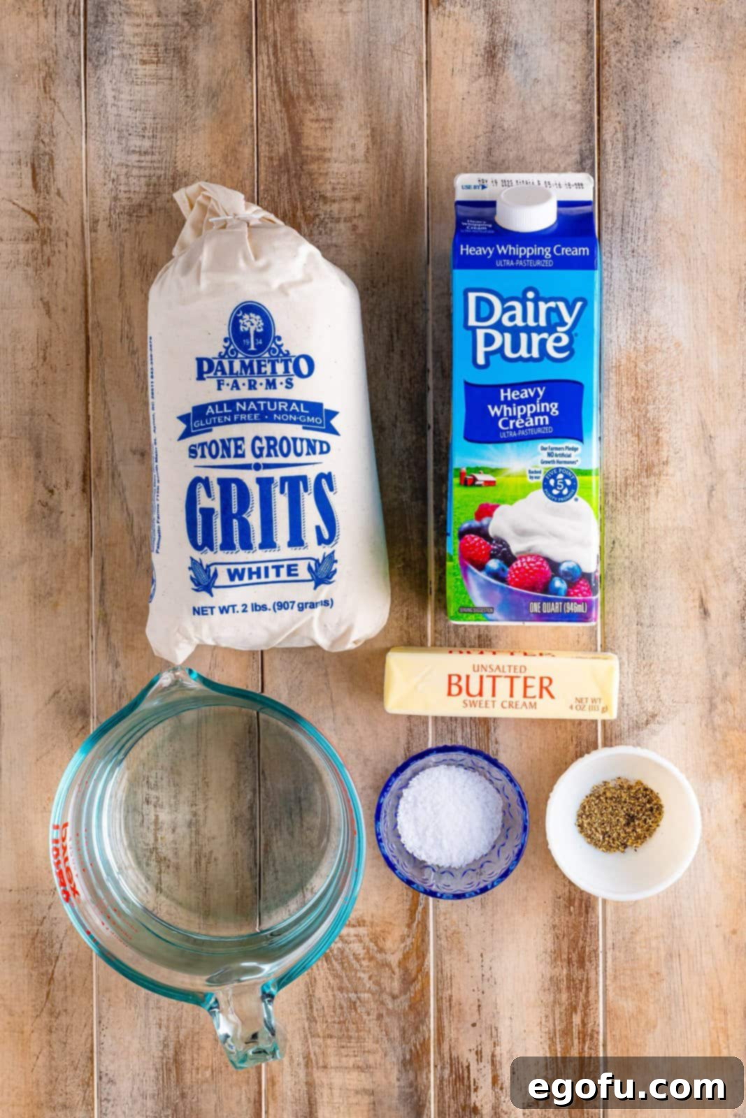 An array of fresh ingredients: stone ground grits, unsalted butter, heavy cream, salt, black pepper, and water, laid out for cooking.