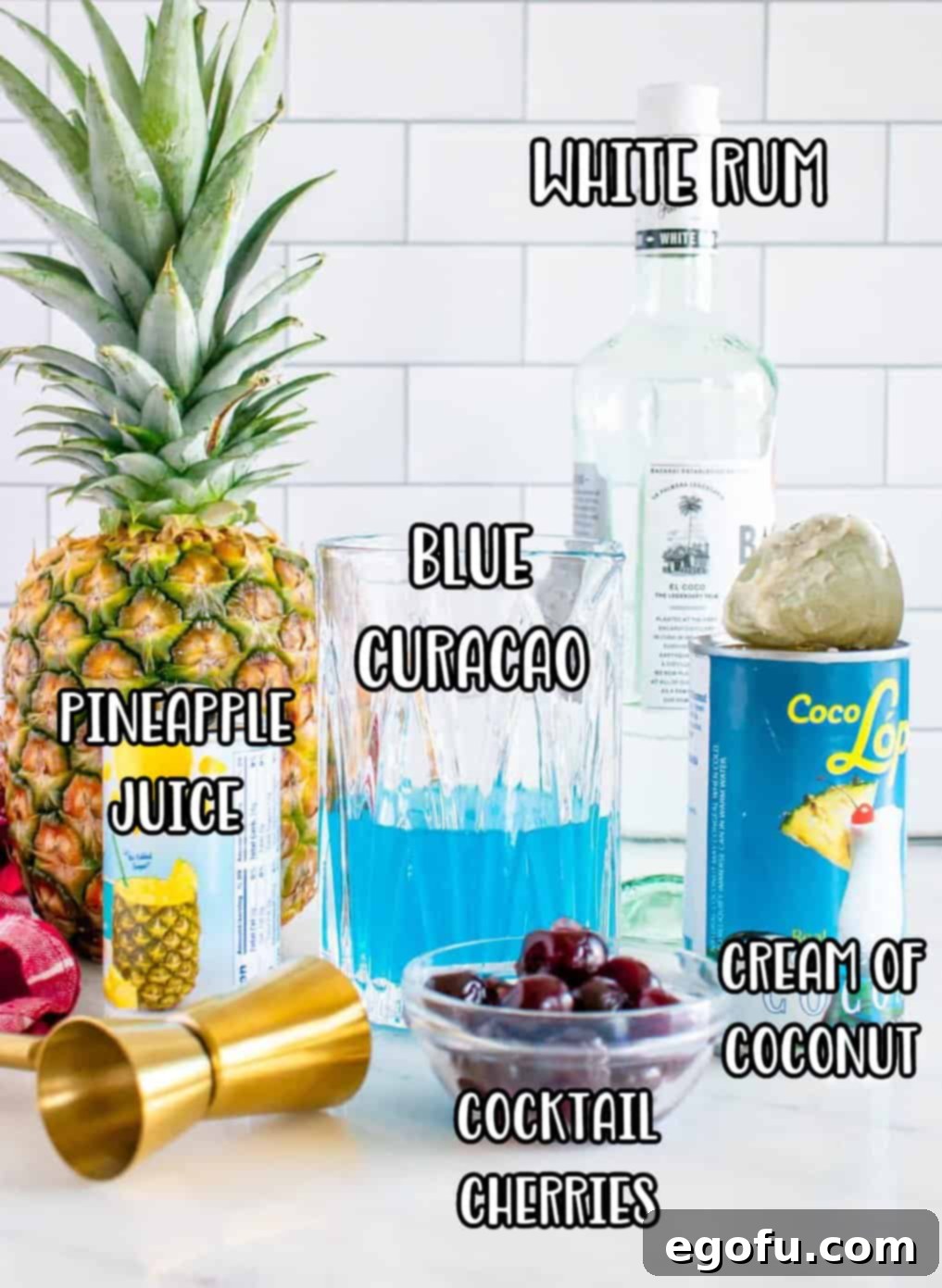 A flat lay showing all ingredients for Blue Hawaiian Cocktail: white rum, blue curaçao, pineapple juice, cream of coconut, cocktail cherries, and pineapple chunks.
