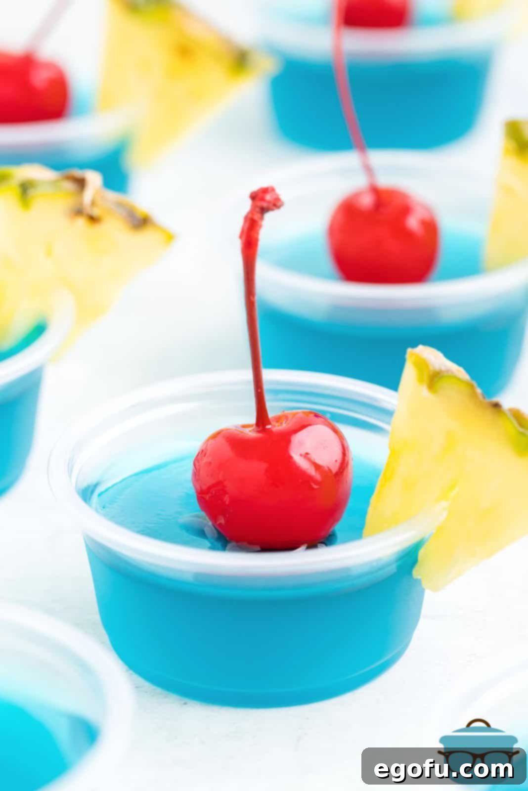 Several Blue Hawaii Jell-O Shots, garnished with fresh pineapple wedges and maraschino cherries, presented on a white serving tray.
