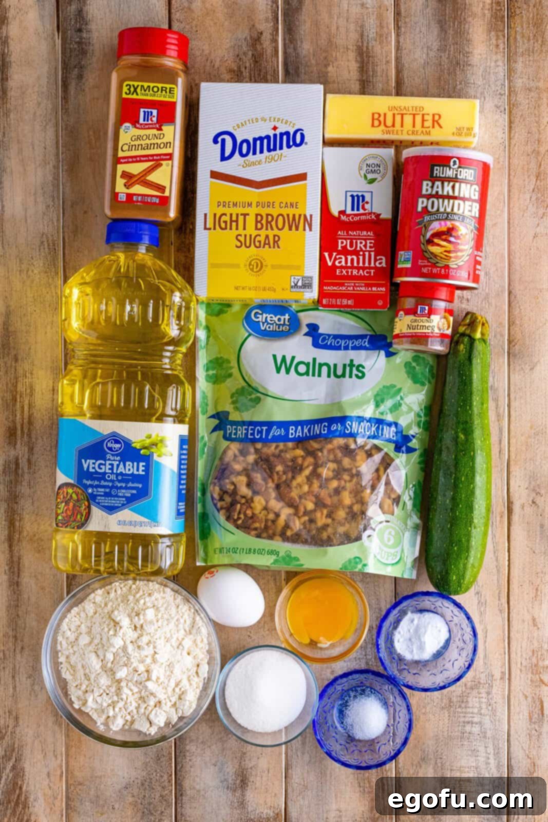 A flat lay photograph showcasing all the fresh ingredients needed for baking zucchini bread: all-purpose flour, salt, baking powder, baking soda, ground cinnamon, nutmeg, a large egg, egg yolk, vegetable oil, unsalted butter, light brown sugar, granulated sugar, vanilla extract, shredded zucchini, and chopped walnuts.