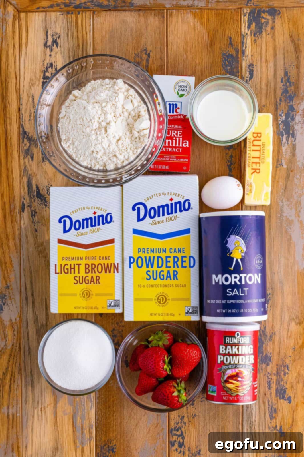 A beautifully arranged flat lay of all the essential ingredients: flour, butter, brown sugar, fresh strawberries, baking powder, salt, granulated sugar, an egg, vanilla extract, milk, and powdered sugar, ready for baking.