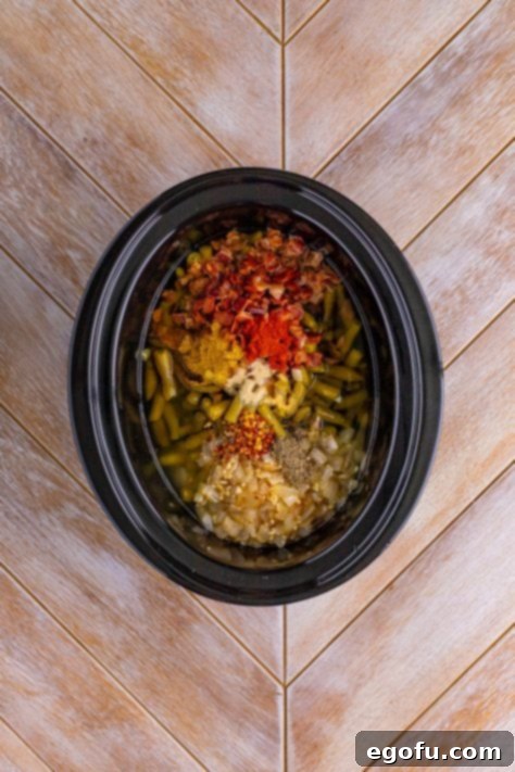 A slow cooker with green beans, half of the bacon, the cooked onions and garlic, chicken stock, butter, seasoned salt, black pepper, paprika, garlic powder, onion powder, and chili flakes.