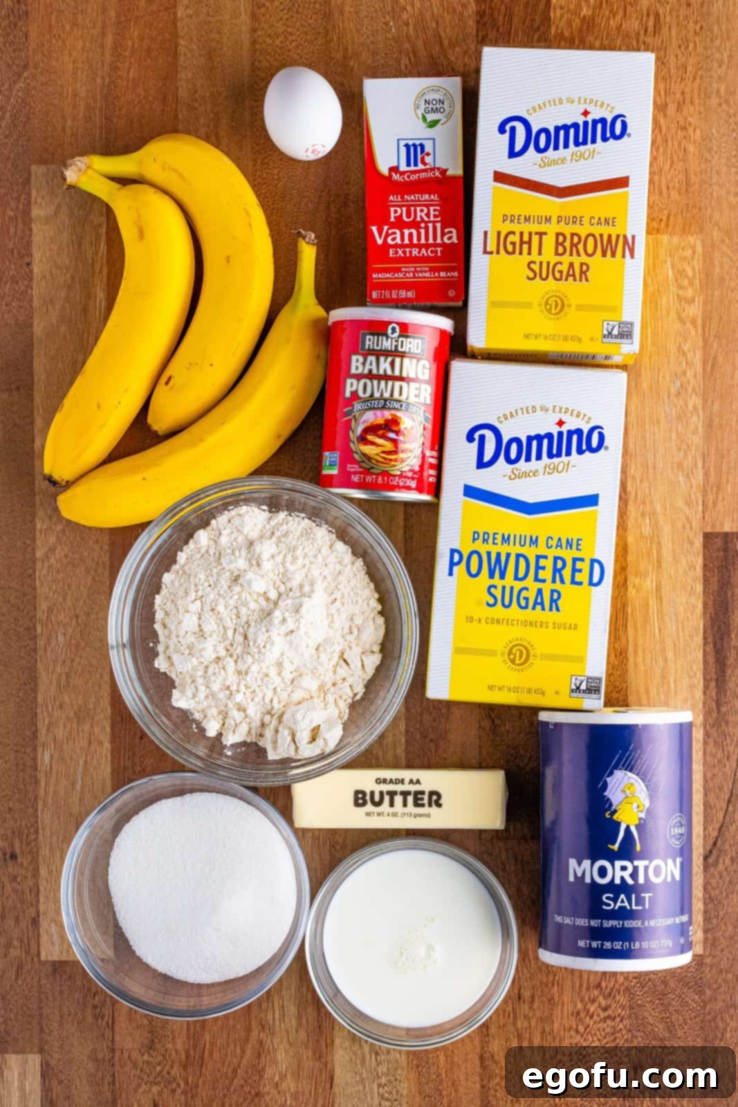 A flat lay photograph showcasing the key ingredients for banana upside down cake: bananas, light brown sugar, granulated sugar, butter, salt, all-purpose flour, vanilla extract, a whole egg, and baking powder.