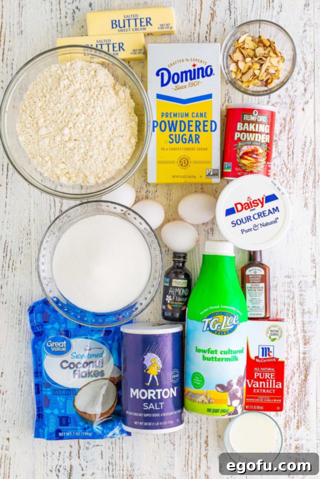 Southern Crunch Delight 4 A vibrant flat lay of all the essential ingredients for Louisiana Crunch Cake, including flour, butter, sugars, eggs, sour cream, extracts, buttermilk, coconut flakes, and sliced almonds.