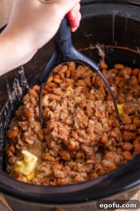A potato masher mashing the beans in a slow cooker.