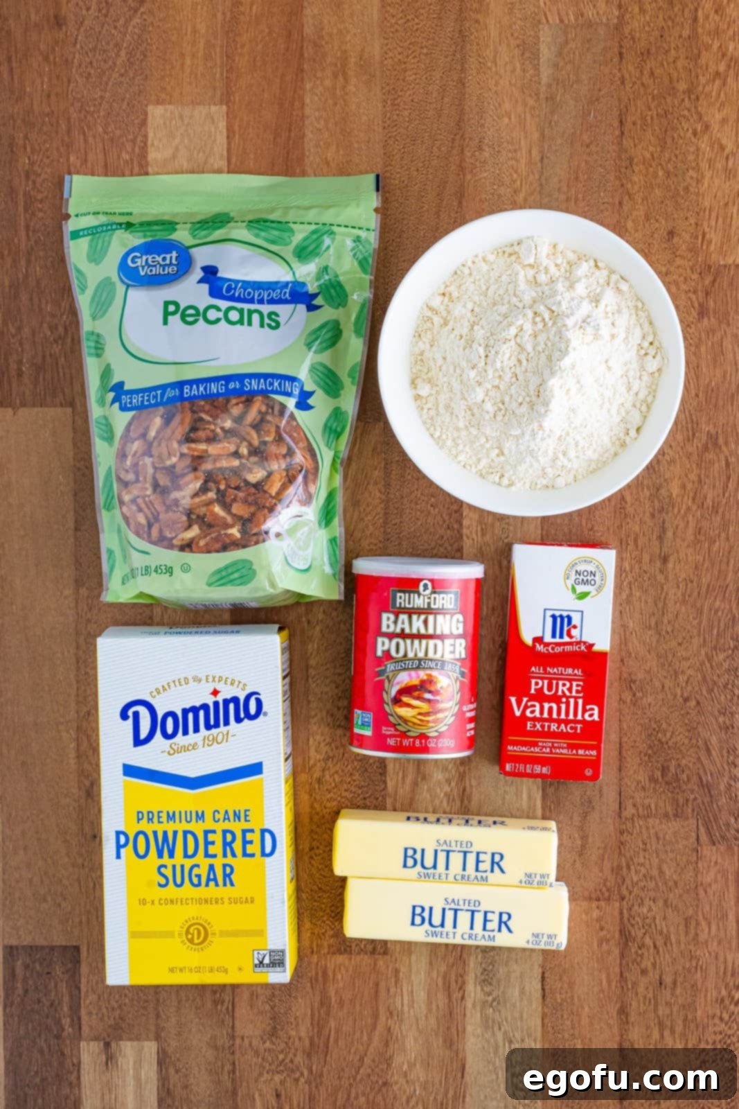 A beautifully composed flat lay showing all the fresh ingredients required for making Pecan Sandies: whole pecans, powdered sugar, softened butter sticks, vanilla extract bottle, baking powder, and a bowl of all-purpose flour.
