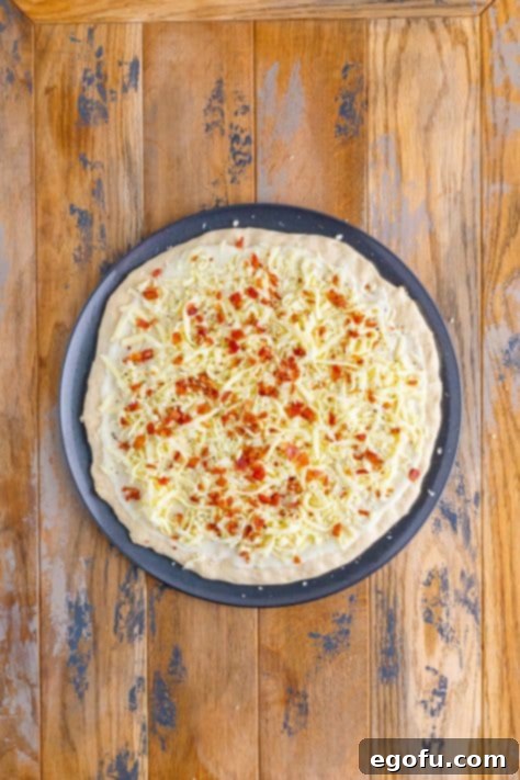 A pizza pan with dough, white sauce, cheese and bacon.