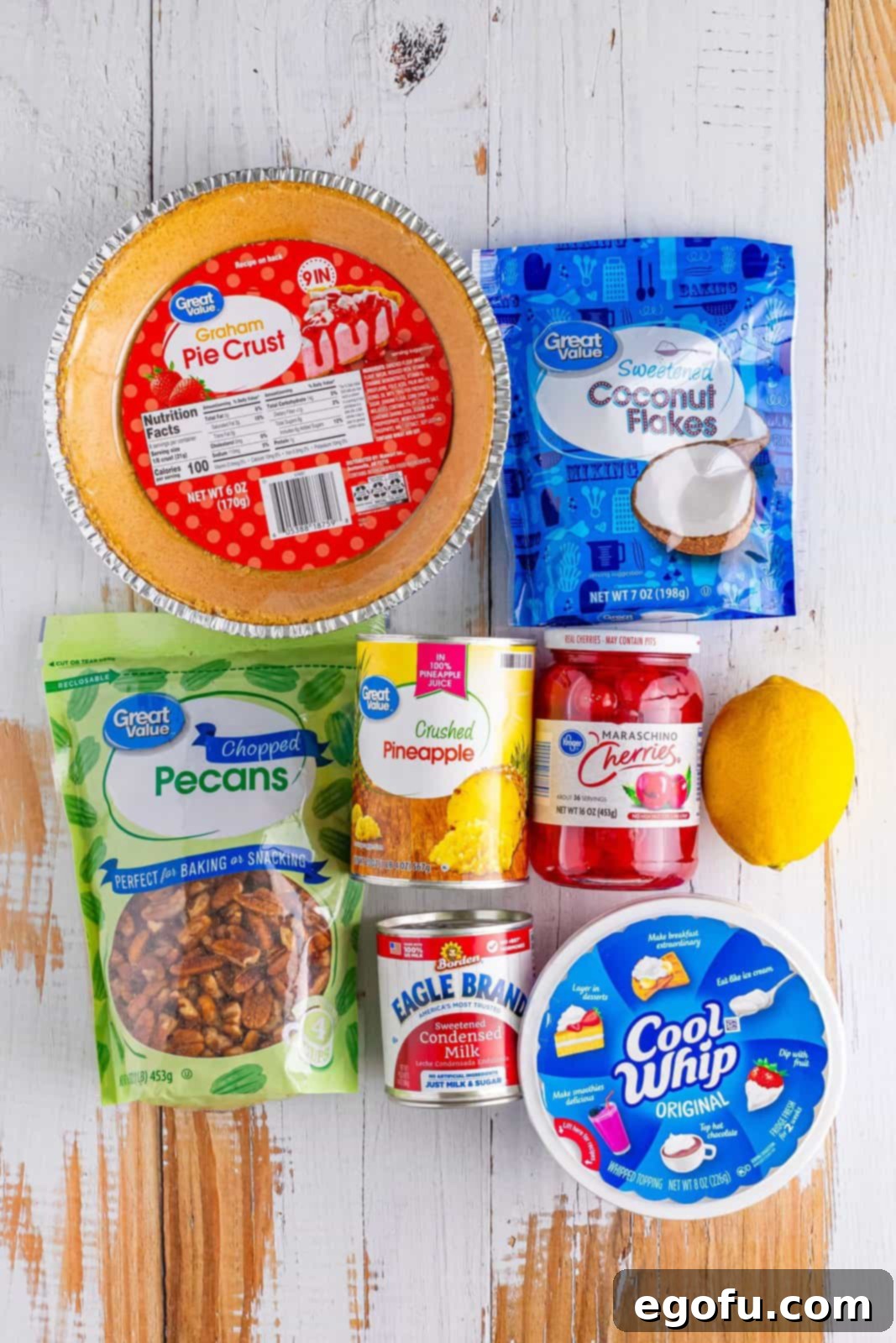 A flat lay photograph showcasing all the fresh and canned ingredients needed for Millionaire Pie: graham cracker crust, Cool Whip, pecans, maraschino cherries, pineapple chunks, a whole lemon, sweetened condensed milk, and coconut flakes, neatly arranged on a white surface.