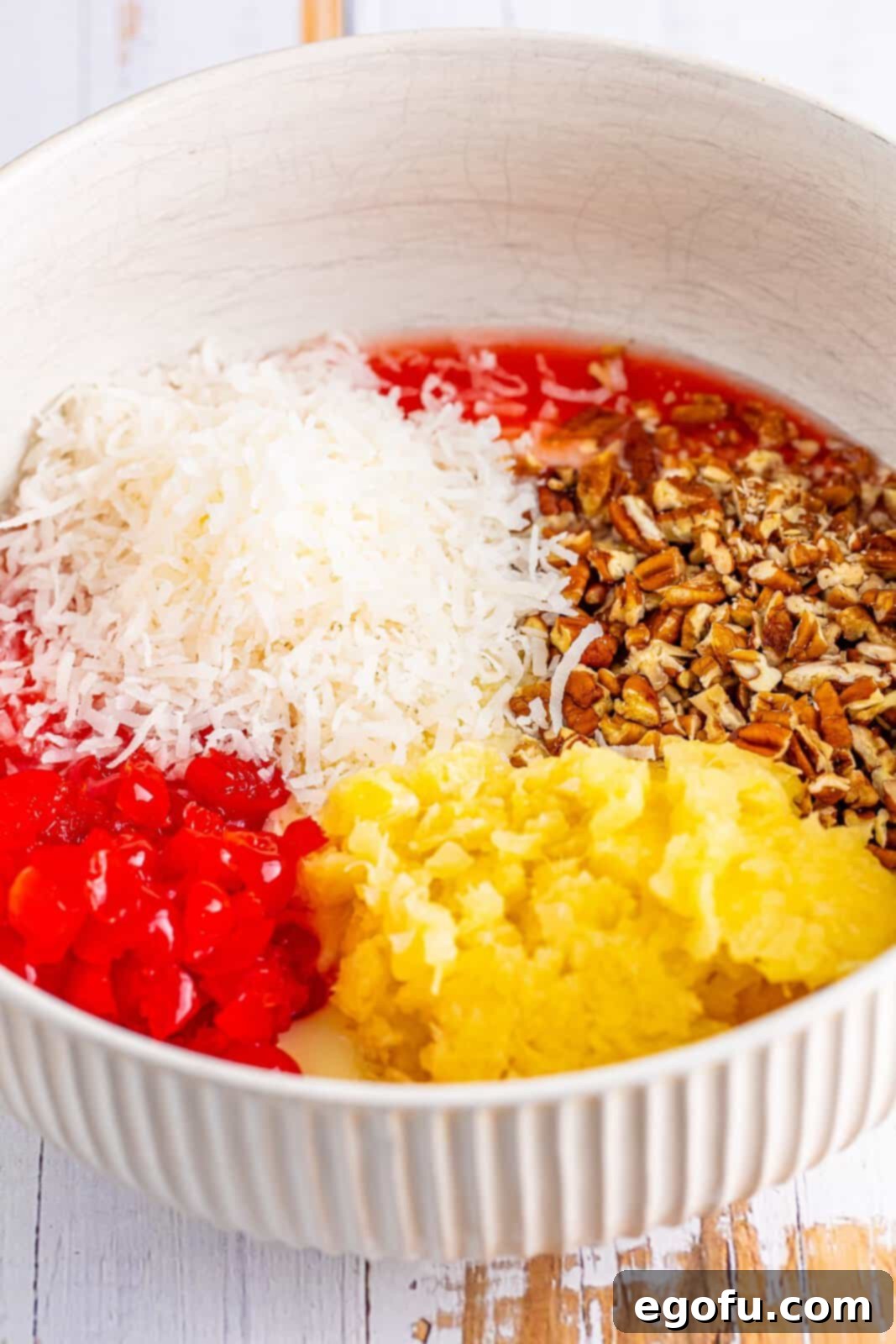 Close-up shot of a large mixing bowl filled with the colorful Millionaire Pie base ingredients: drained pineapple, condensed milk, coconut flakes, maraschino cherries, pecans, lemon juice, and maraschino cherry juice, all stirred together.