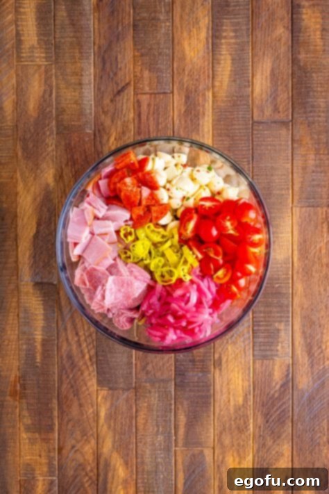 A large bowl of cooked rotini, cut up mozzarella balls, sliced pepperoncini peppers, cherry tomatoes, pickled red onion, salami, ham and pepperoni.