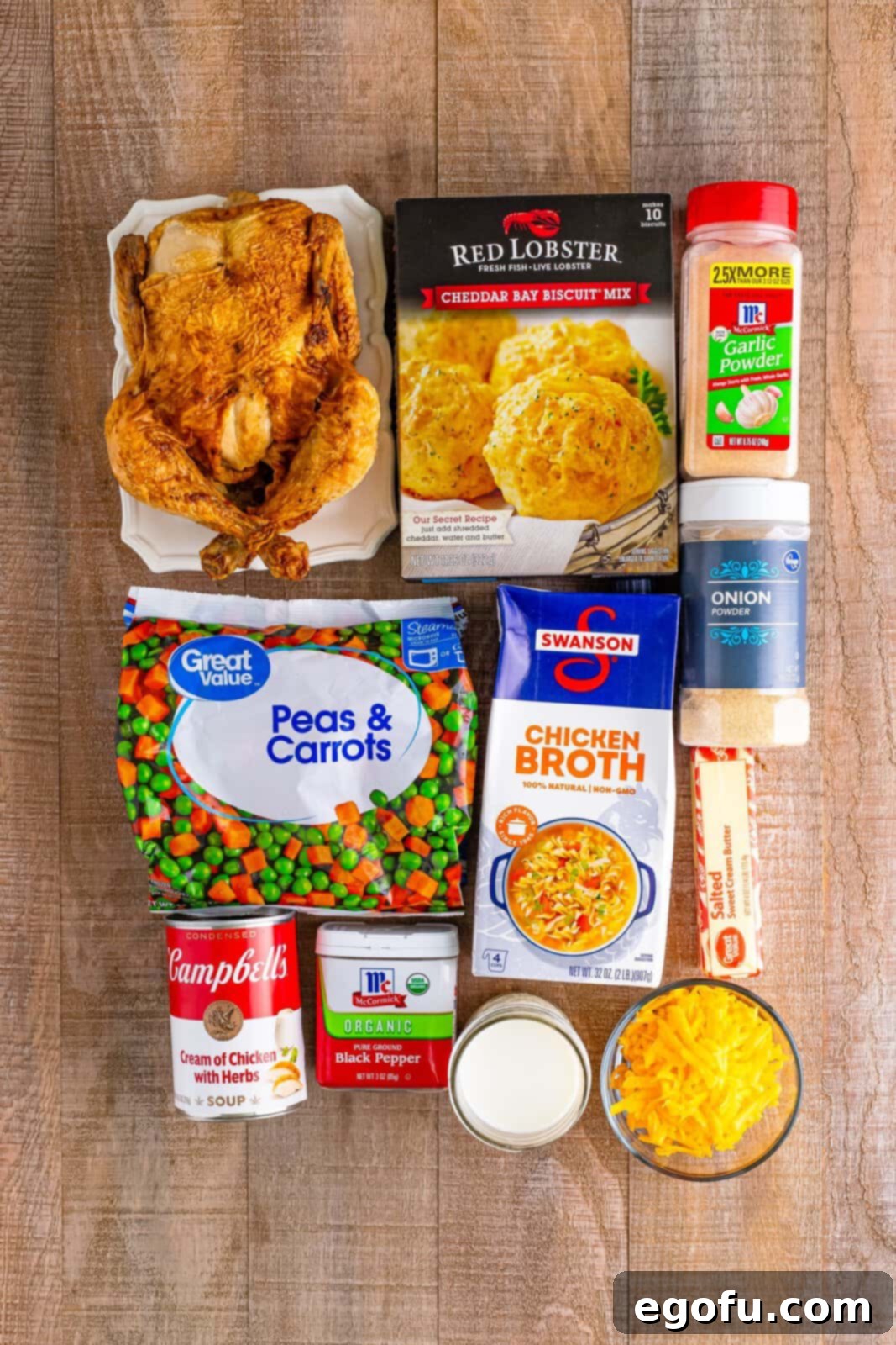 A vibrant flat lay of all the essential ingredients for Chicken Cobbler: butter, rotisserie chicken, frozen peas and carrots, cream of chicken soup, chicken broth, various seasonings, Cheddar Bay Biscuit Mix, milk, and shredded cheddar cheese.