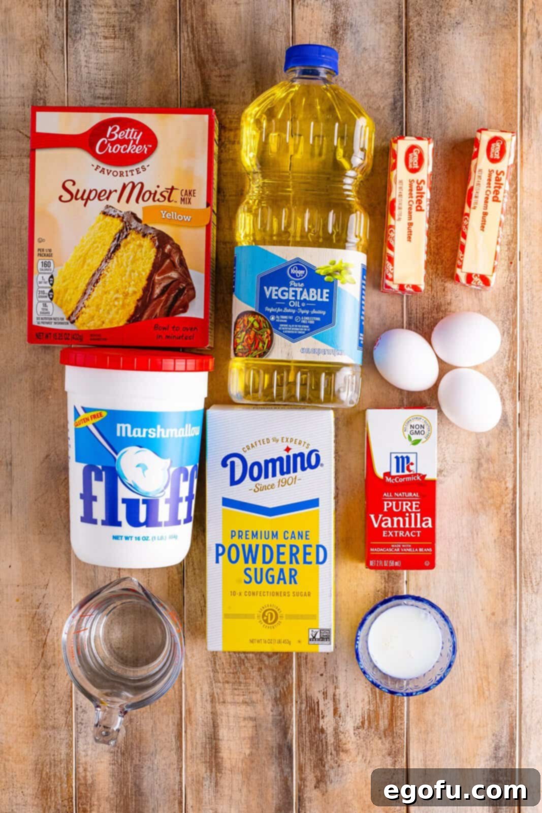A flat lay of all the essential ingredients for Twinkie Poke Cake: yellow cake mix, oil, eggs, water, softened butter, marshmallow fluff, powdered sugar, whole milk, and vanilla extract.