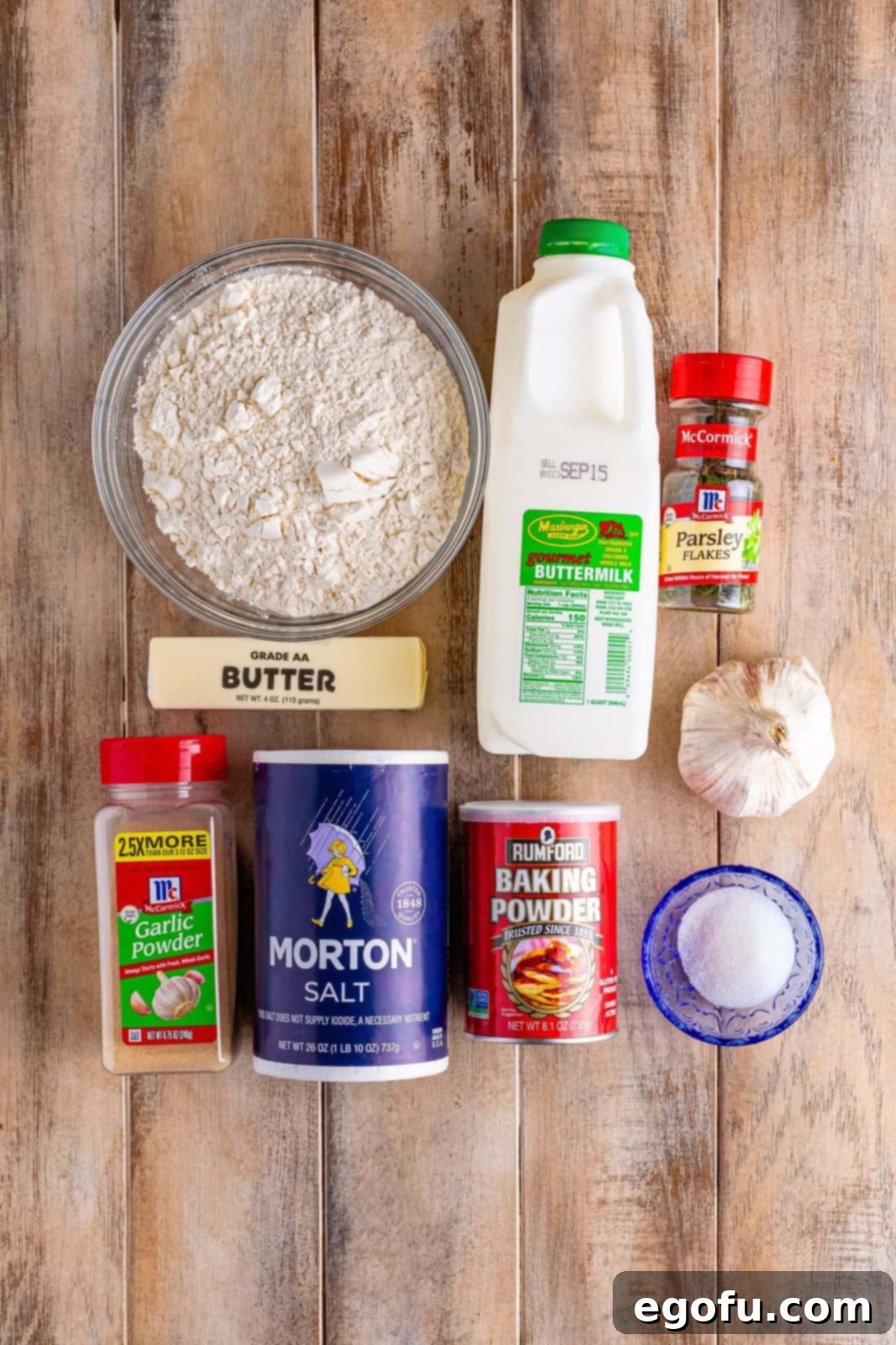 Garlic Butter Bath Biscuits 4 A beautifully arranged flat lay showing all the raw ingredients for Garlic Butter Swim Biscuits: all-purpose flour, sugar, baking powder, salt, butter, garlic powder, buttermilk, fresh garlic cloves, and dried parsley flakes.
