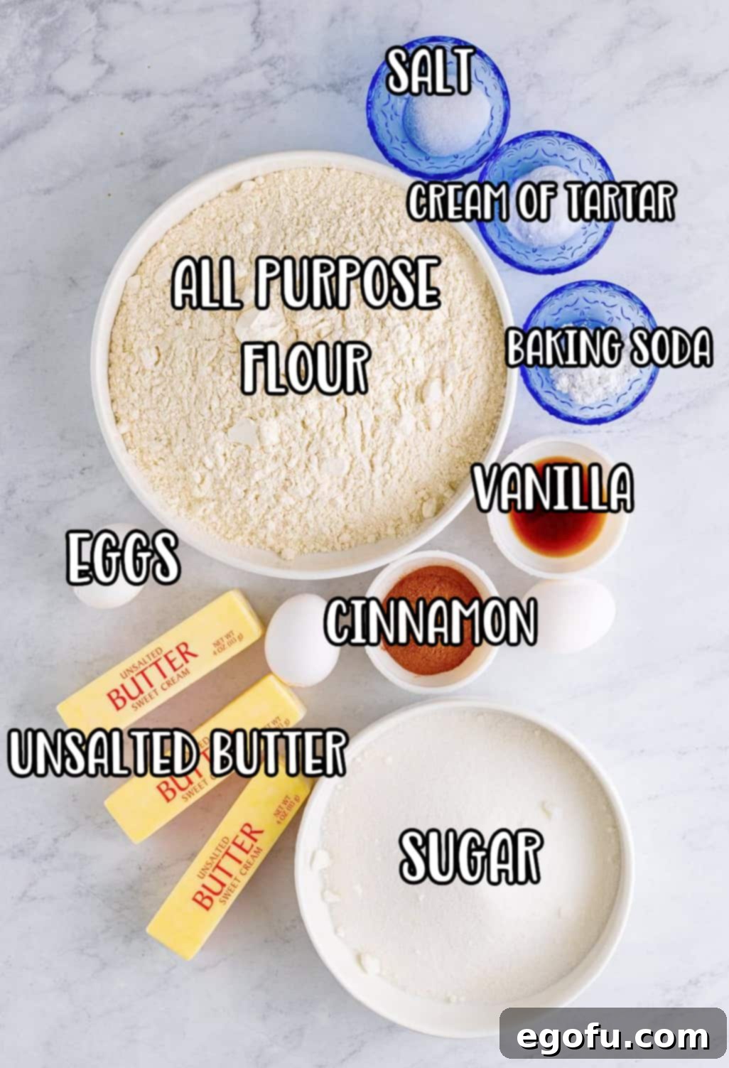 A flat lay photograph displaying all the essential ingredients for Crumbl Snickerdoodles: all-purpose flour, cream of tartar, baking soda, salt, unsalted butter, granulated sugar, large eggs, vanilla extract, and ground cinnamon.