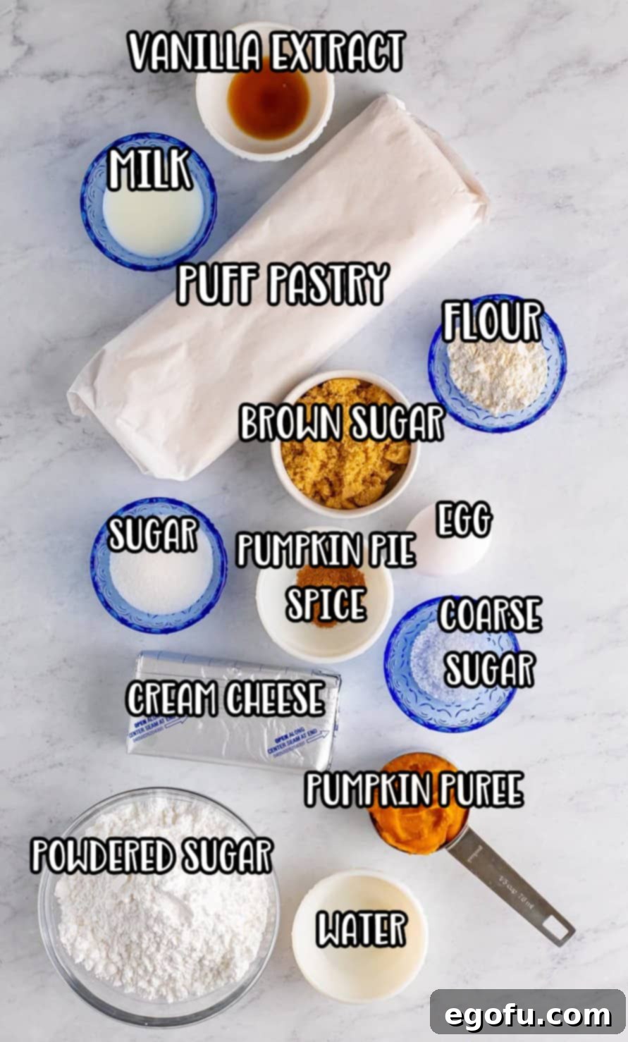 An array of ingredients laid out, including cream cheese, vanilla extract, sugar, pumpkin puree, light brown sugar, all purpose flour, pumpkin pie spice, puff pastry, egg, water, coarse sugar, powdered sugar, and milk.
