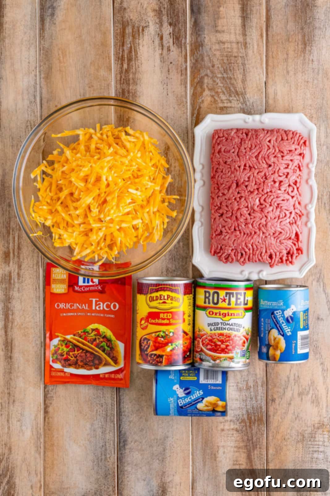 A flat lay photograph showcasing all the raw ingredients: ground beef, shredded cheese, taco seasoning, enchilada sauce, diced tomatoes with green chilies, and canned biscuit dough, neatly arranged on a rustic surface.