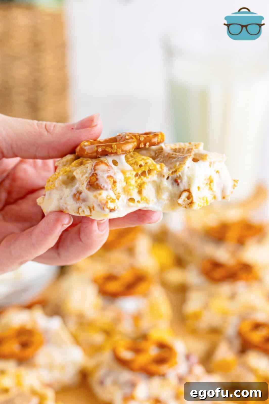 A hand holding a perfectly square Chex Mix Marshmallow Treat, showcasing its gooey texture and visible pretzel pieces.