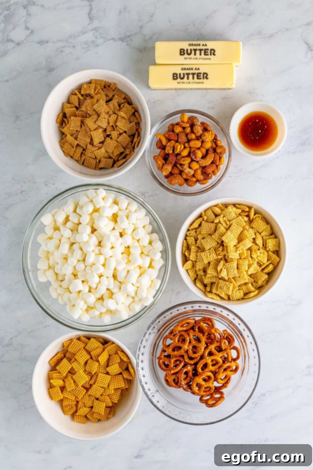 An organized arrangement of all the ingredients: rice chex, wheat chex, corn chex, butter, marshmallows, vanilla extract, mixed nuts, and pretzels laid out for baking.
