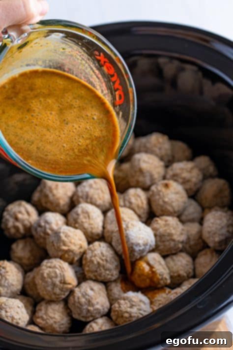 A glass measuring cup with au jus gravy mix, ranch dressing mix, water, and pepperoncini juice being poured over meatballs in a Crock Pot.
