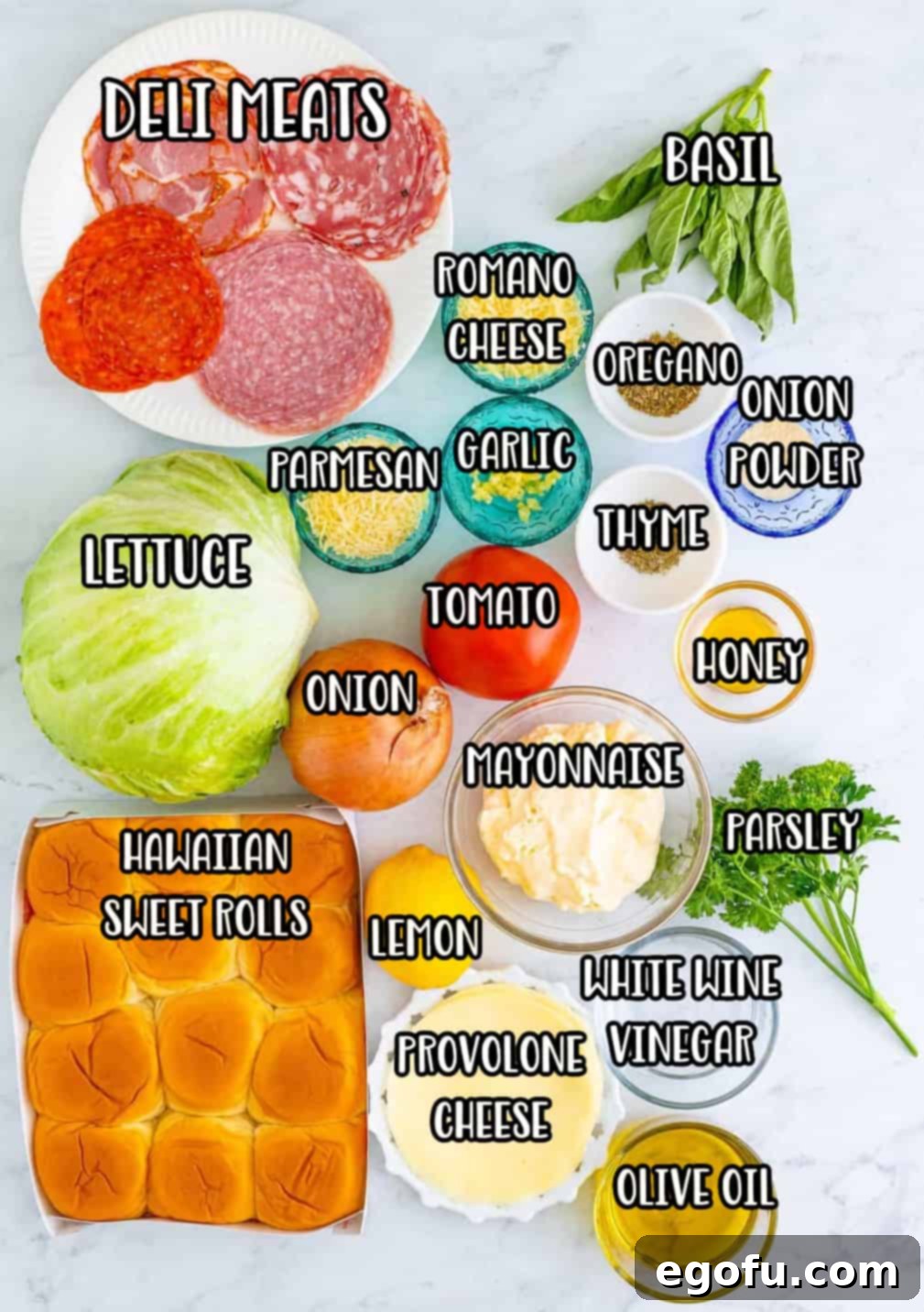 A carefully arranged flat lay of all the fresh and dry ingredients required for Italian Grinder Sliders, including mayonnaise, olive oil, vinegars, cheeses, lemon, garlic, honey, basil, parsley, oregano, onion powder, thyme, tomatoes, sweet onion, lettuce, Hawaiian rolls, provolone cheese, pepperoni, salami, capocollo, and soppressata.