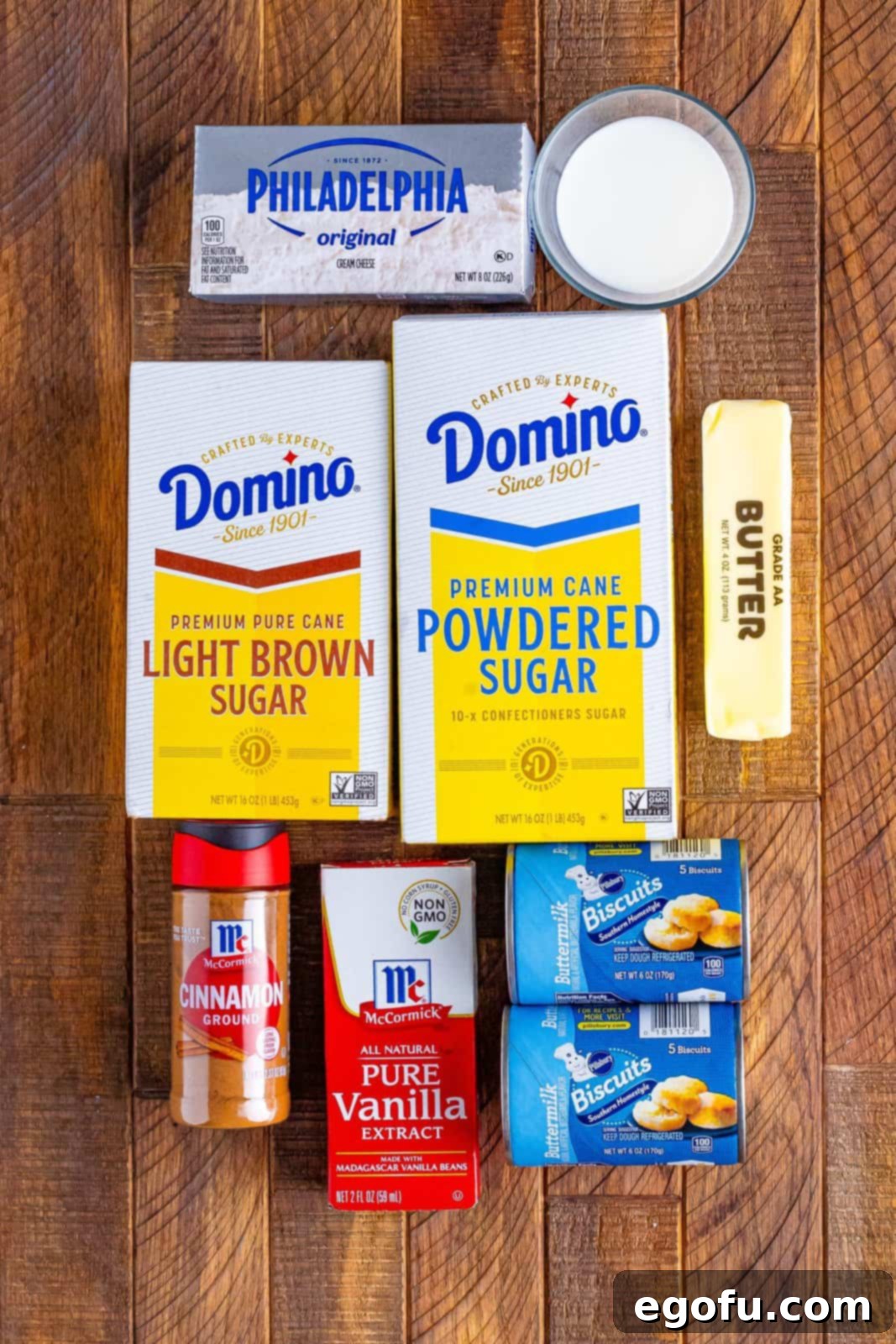 A flat lay of all the essential ingredients for Cinnamon Roll Bubble Up: light brown sugar, salted butter, ground cinnamon, vanilla extract, two cans of biscuits, a block of cream cheese, powdered sugar, and a glass of milk.