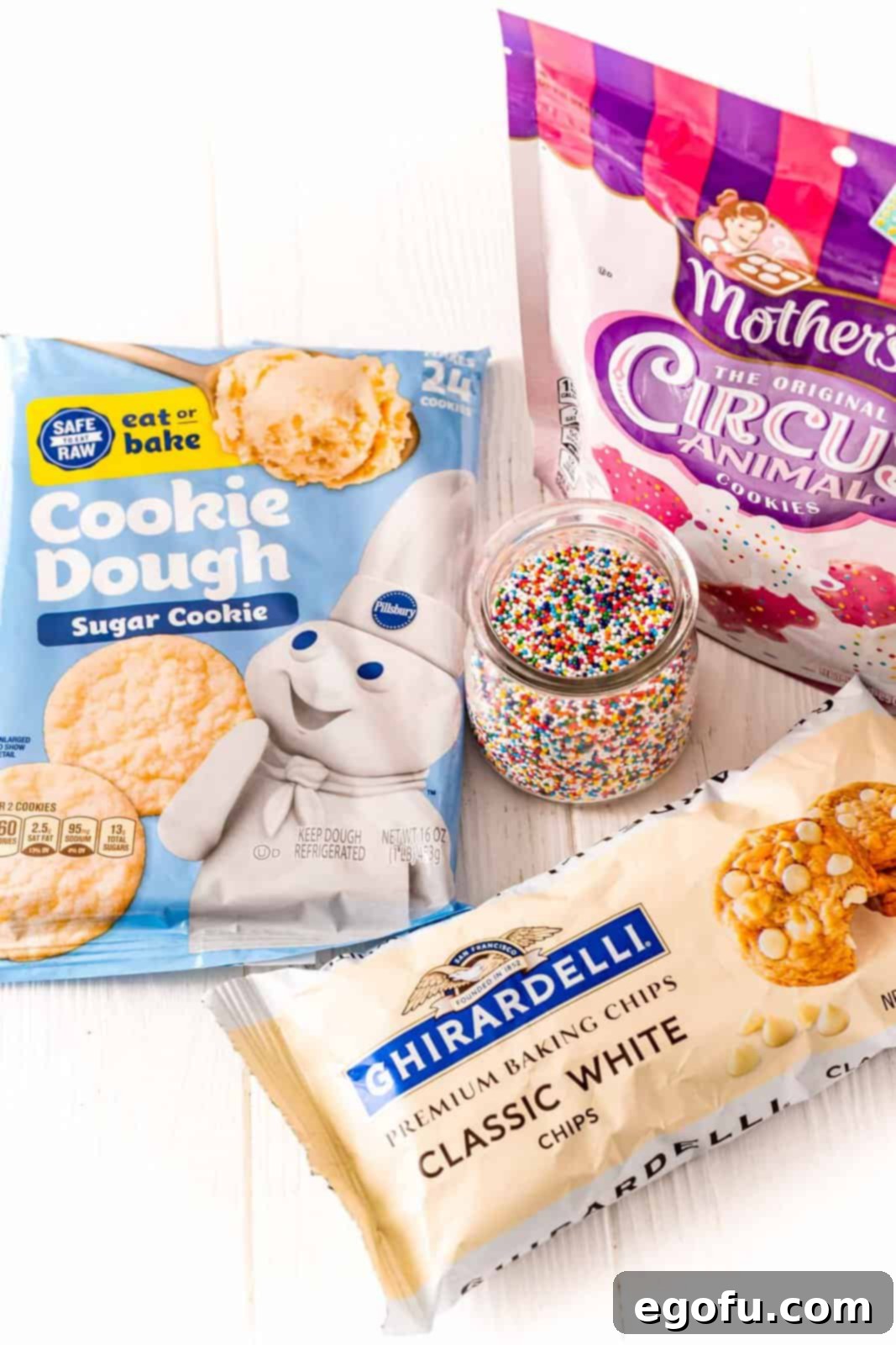 The four main ingredients for Circus Animal Cookies laid out: frosted animal cookies, refrigerated sugar cookie dough, white chocolate chips, and rainbow nonpareils.