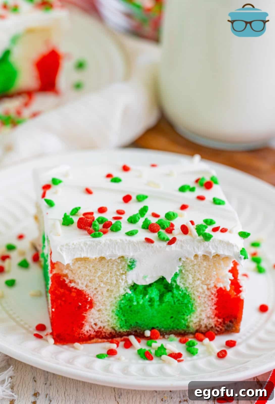 A vibrant slice of Christmas Jell-O Poke Cake on a plate, showcasing distinct red and green layers infused into the moist white cake, topped with fluffy whipped cream and festive sprinkles.