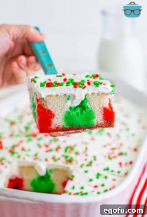 A close-up of a serving utensil holding a perfect slice of Christmas Jell-O Poke Cake, showcasing the vibrant layers and sprinkles, hovering over the rest of the beautifully decorated cake.