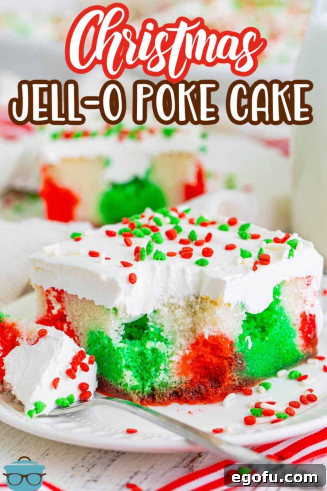 A close-up of a fork holding a bite of Christmas Jell-O Poke Cake, showing the moist cake, gelatin layers, and whipped topping, with a slice of cake in the background.