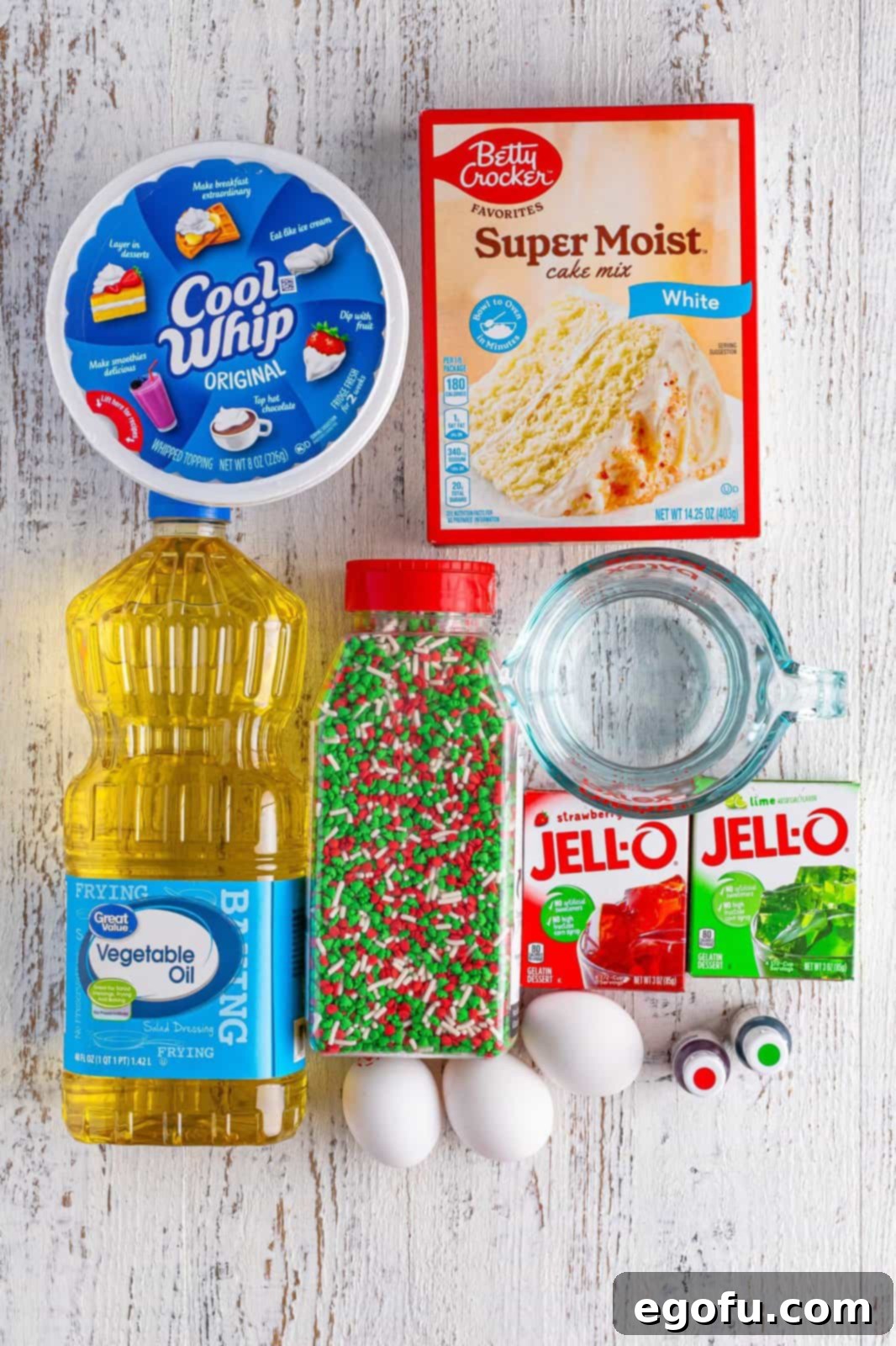 An array of ingredients laid out for Christmas Jell-O Poke Cake, including a box of white cake mix, eggs, vegetable oil, water, red and green Jell-O mixes, a tub of whipped topping, and Christmas-themed sprinkles.