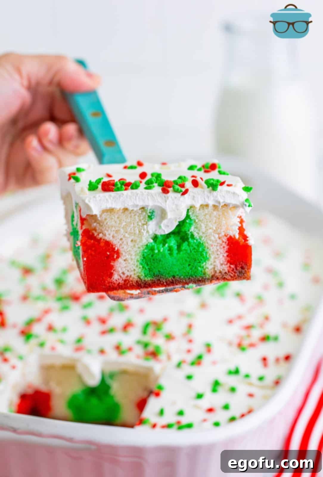 A serving utensil lifts a perfectly sliced piece of Christmas Jell-O Poke Cake, showcasing the festive red and green gelatin layers and fluffy white topping, with the remaining cake in the background.