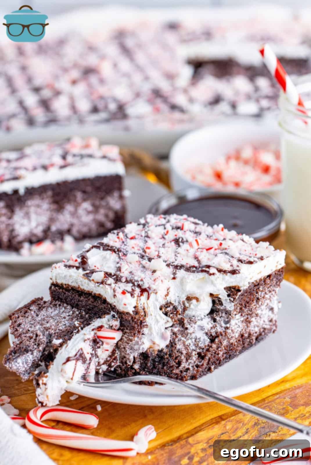 A fork holding a bite of Peppermint Poke Cake next to a slice on a plate, showcasing the chocolate cake, marshmallow filling, whipped topping, and crushed peppermints.