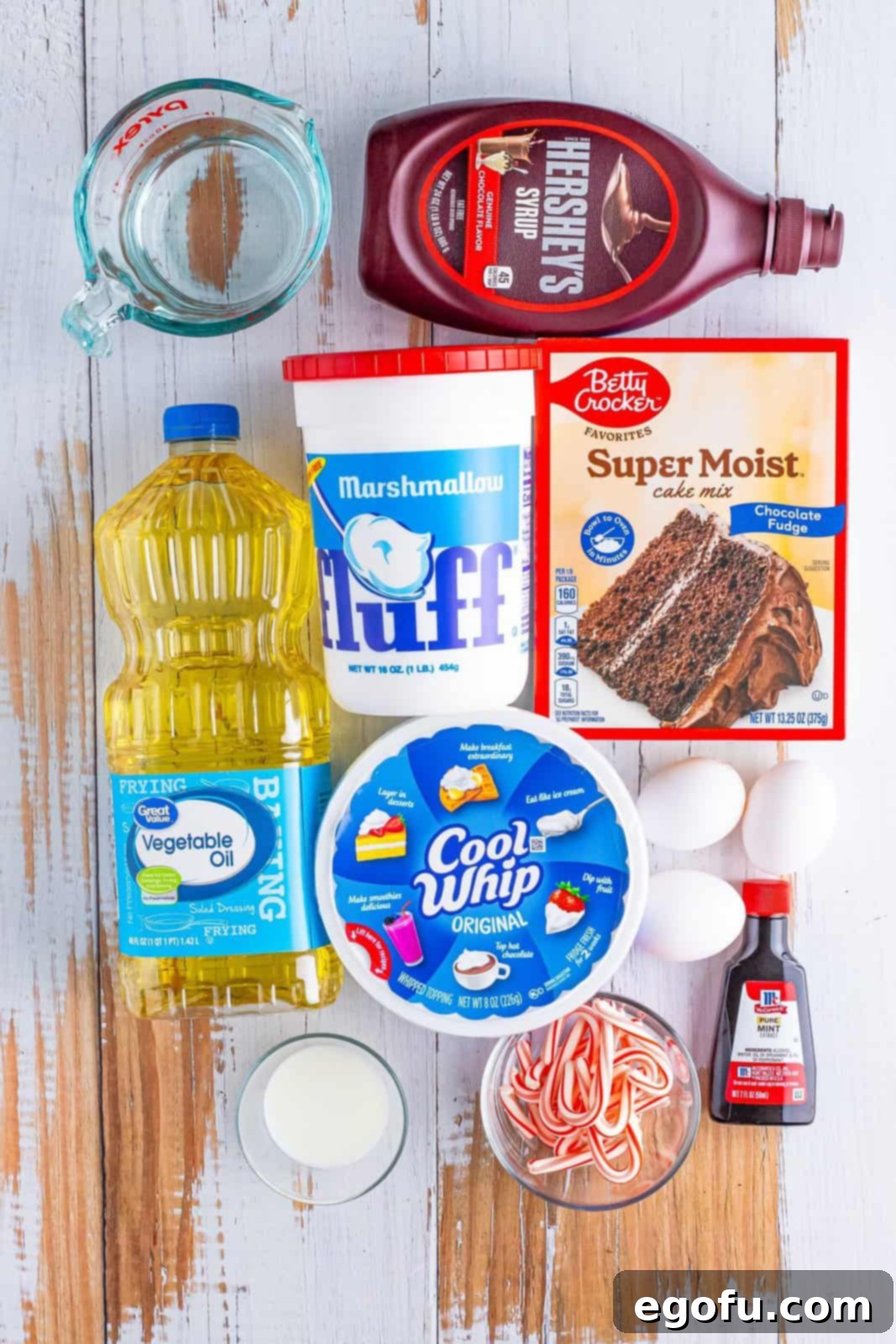 An array of ingredients for Peppermint Poke Cake: Marshmallow fluff, Cool Whip, chocolate cake mix, eggs, milk, vegetable oil, chocolate sauce, mint extract, and crushed candy canes laid out on a surface.
