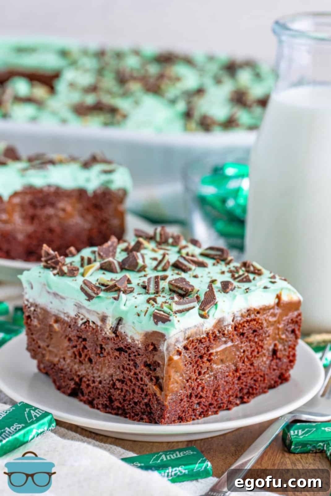 Peppermint Patty Poke Cake 2 A perfectly sliced piece of homemade Mint Chocolate Poke Cake on a pristine white serving plate, with the rest of the cake visible in its baking dish in the background.