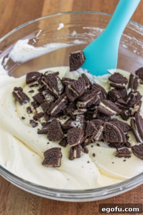Oreo pieces being added to the cream cheese mixture in a mixing bowl.