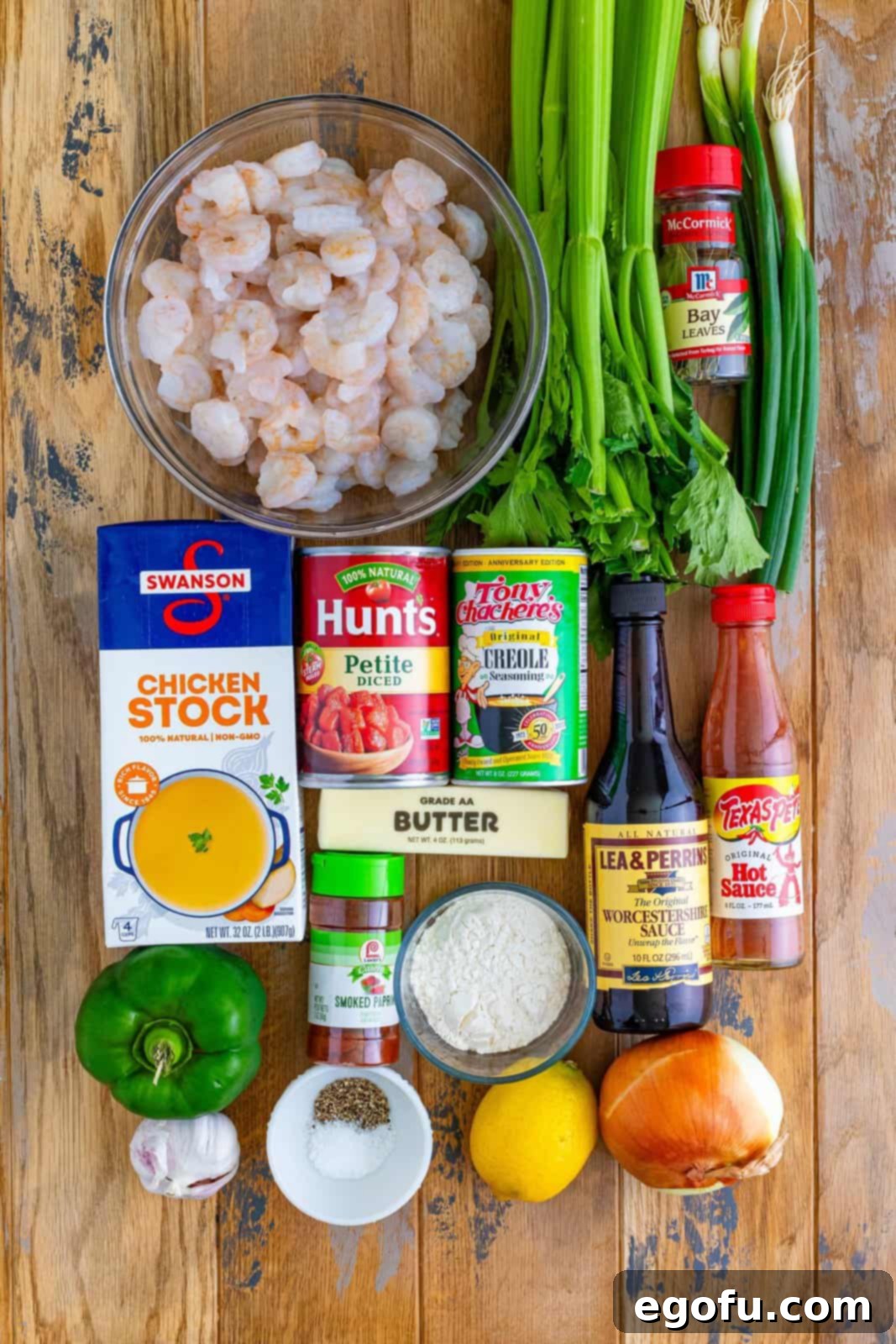 An artfully arranged spread of fresh and vibrant ingredients: raw medium shrimp, thinly sliced celery and green onion, kosher salt, black pepper, a jar of Creole seasoning, a bottle of hot sauce, Worcestershire sauce, a fresh lemon, a can of petite diced tomatoes, minced garlic, all-purpose flour, a stick of butter, green bell pepper, sweet onion, chicken stock, a bay leaf, and smoked paprika. All components are meticulously laid out on a rustic surface, signaling their readiness to be transformed into a delicious Shrimp Étouffée.