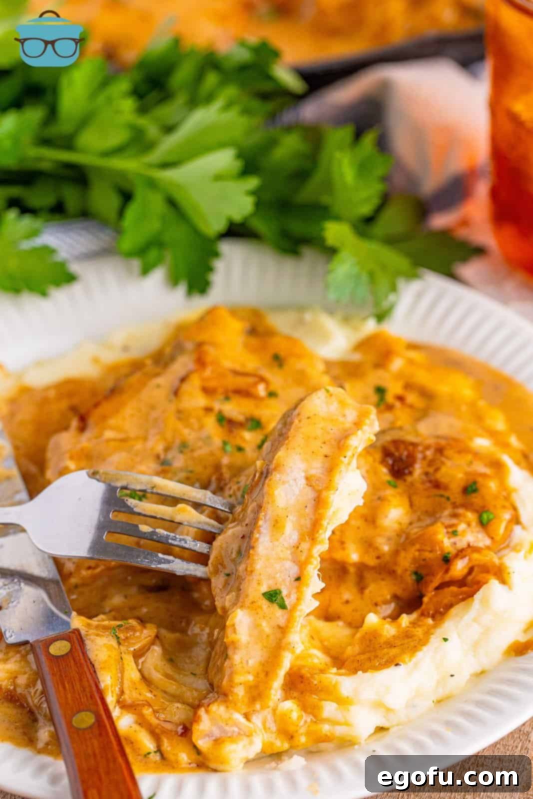 A fork holding a piece of a Smothered Pork Chop above a whole piece on mashed potatoes.