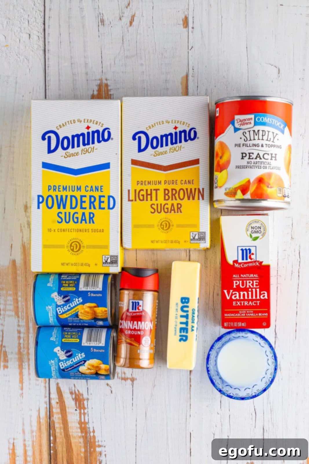A flat lay photograph displaying all the ingredients needed for Peach Dumpling Bubble Up: light brown sugar, powdered sugar, biscuit dough, butter, cinnamon, vanilla extract, peach pie filling, and milk.