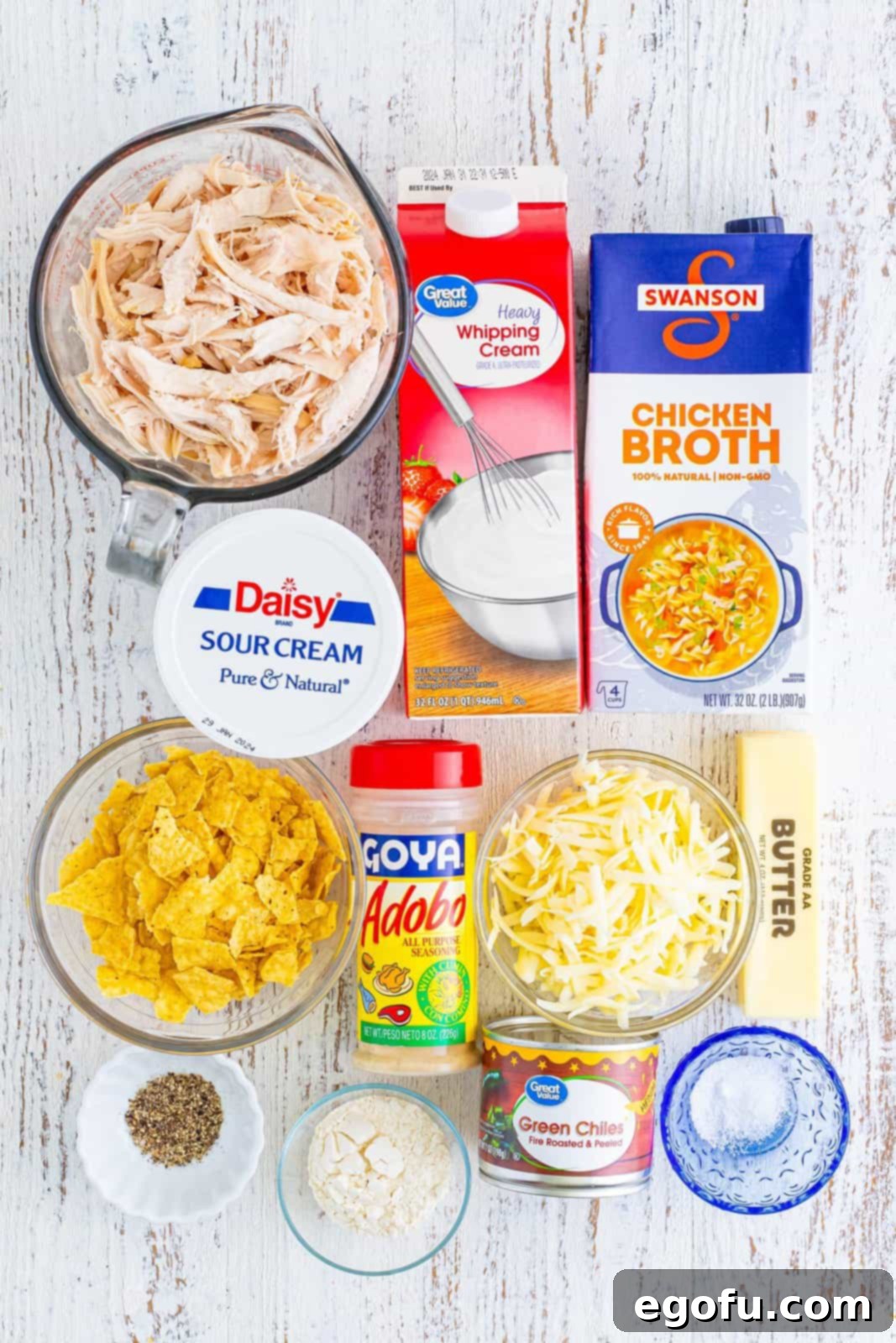 A flat lay of all the fresh ingredients needed to make White Chicken Enchilada Soup: salted butter, flour, adobo seasoning, shredded Monterey Jack cheese, sour cream, black pepper, chicken broth, heavy cream, shredded chicken, and diced green chiles.