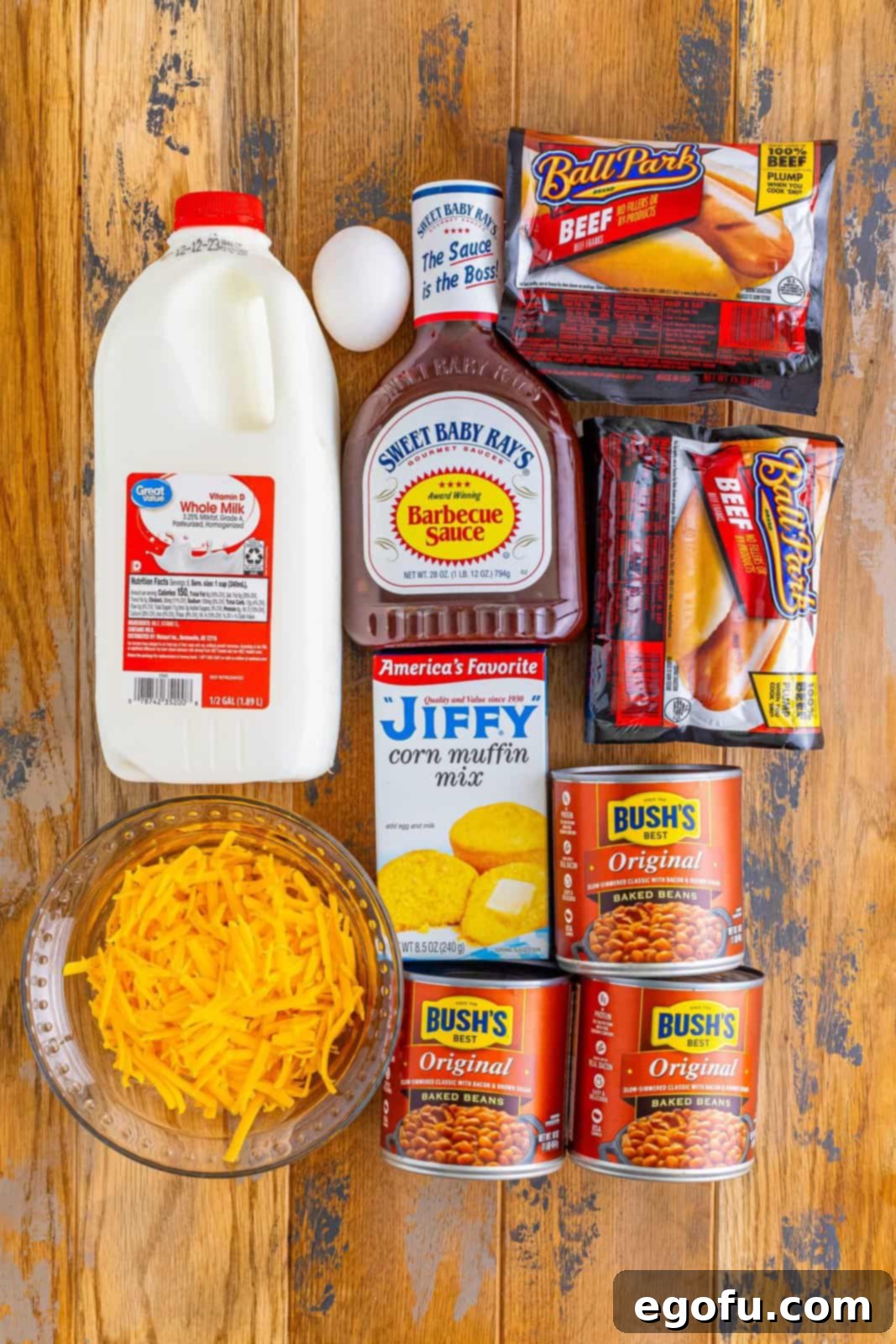 A vibrant flat lay showcasing all the essential ingredients: milk, corn muffin mix, shredded cheese, BBQ sauce, canned baked beans, hot dogs, and eggs.