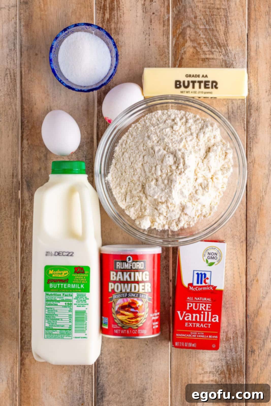 Individual ingredients for buttermilk waffles artfully arranged on a surface: fresh buttermilk, a bowl of all-purpose flour, butter sticks, vanilla extract bottle, baking powder, eggs, and granulated sugar. All components are ready for the mixing process.