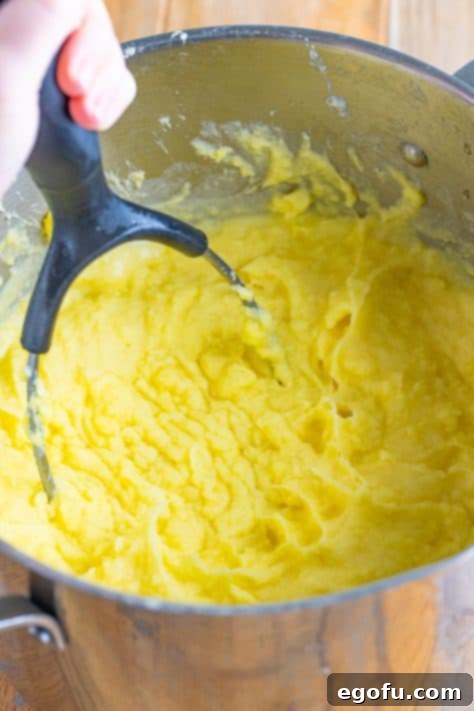 Velvety Stovetop Mashed Potatoes 9 A potato masher actively mashing tender potatoes in a large pot, with warm liquid being incorporated for perfect creaminess.