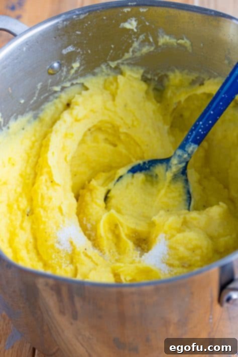 Velvety Stovetop Mashed Potatoes 10 A blue serving spoon mixing freshly ground black pepper and a pinch of salt into a pot of creamy, freshly mashed potatoes.