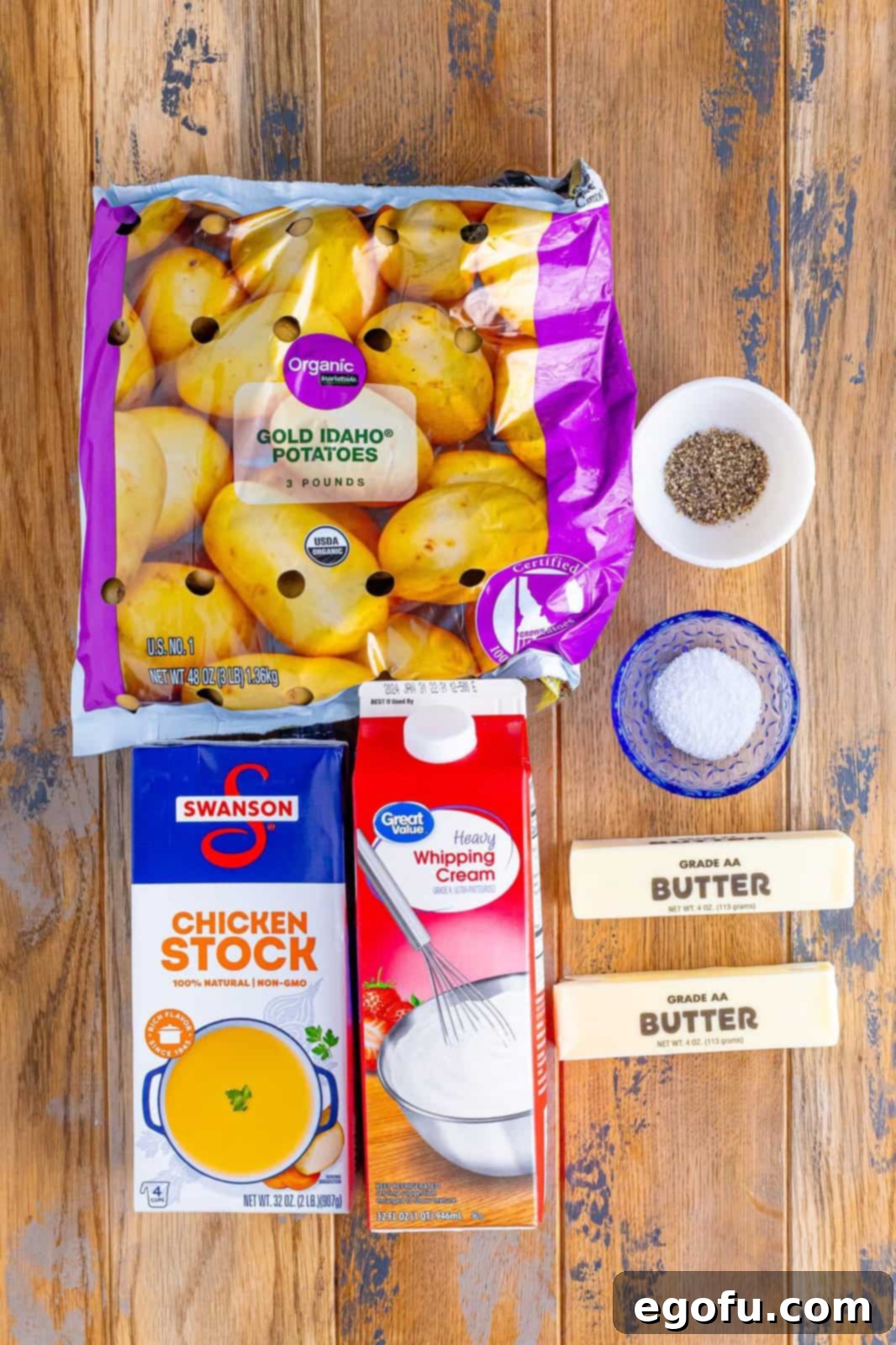 Velvety Stovetop Mashed Potatoes 4 A flat lay photograph displaying fresh Yukon Gold potatoes, a carton of chicken stock, a stick of butter, a small pitcher of heavy whipping cream, and shakers of salt and black pepper—all the essential ingredients for perfect mashed potatoes.