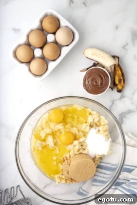 A glass mixing bowl containing the wet ingredients for muffins: melted butter, mashed bananas, both sugars, eggs, and vanilla extract.